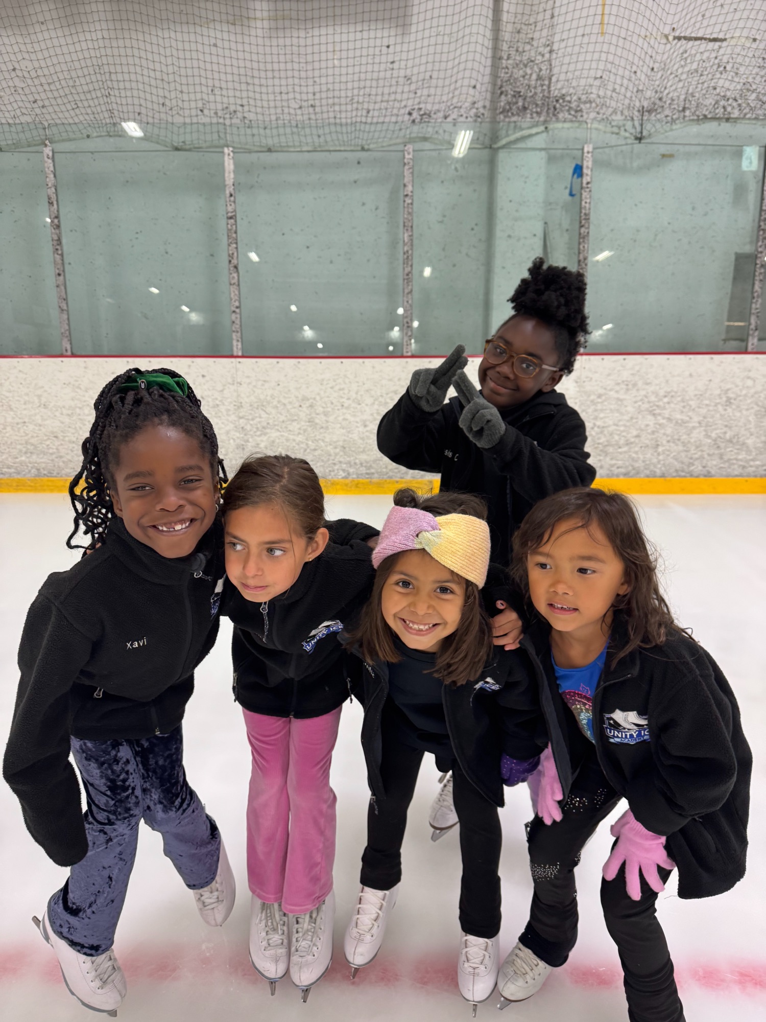 Five smiling young skaters in Unity Ice Academy jackets huddle together on the ice.