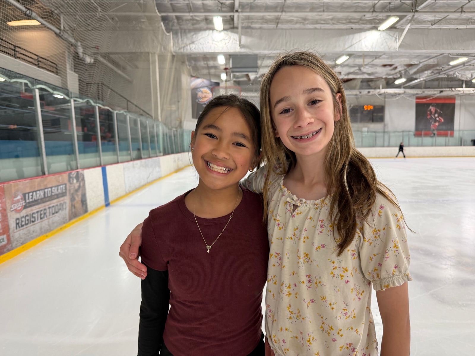 Two smiling young skaters stand arm in arm at the rink.