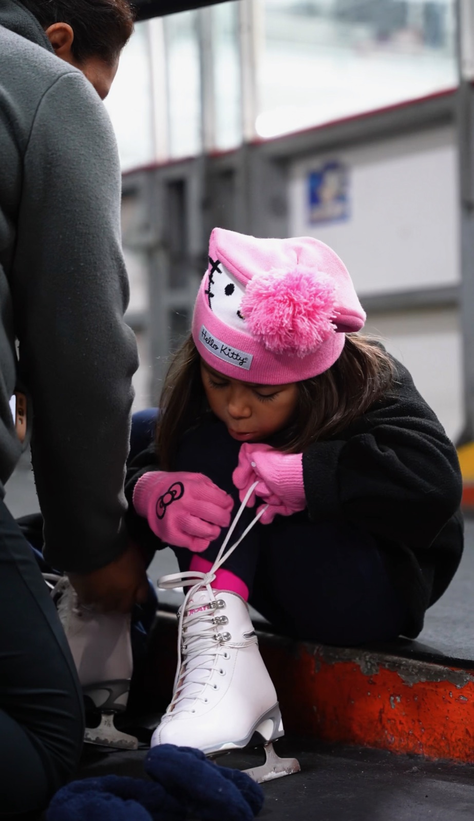 A young skater in a pink hat and gloves laces up her white figure skates while a coach helps.