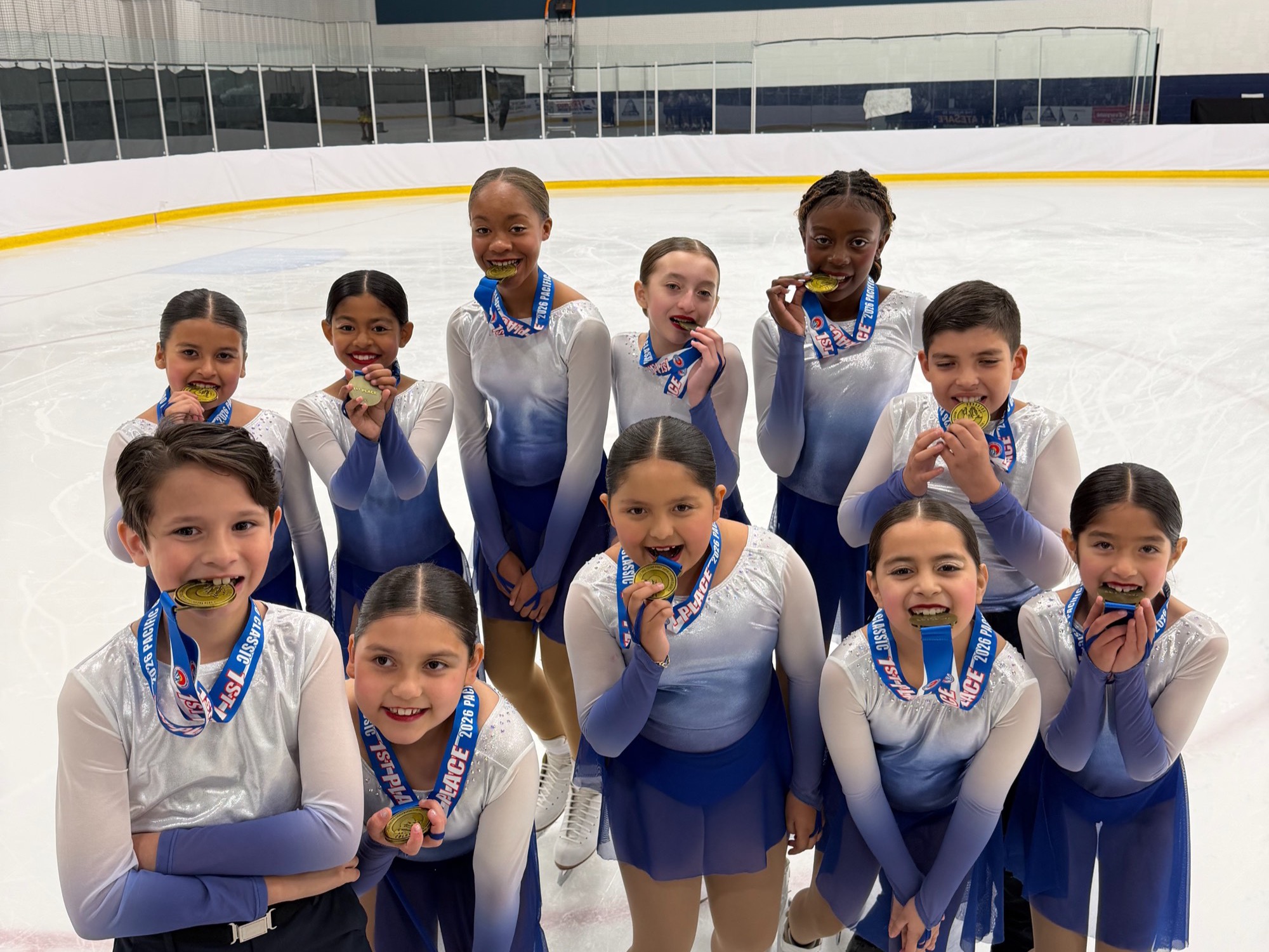 The synchronized skating team in matching blue and white dresses bites their gold medals on the ice.