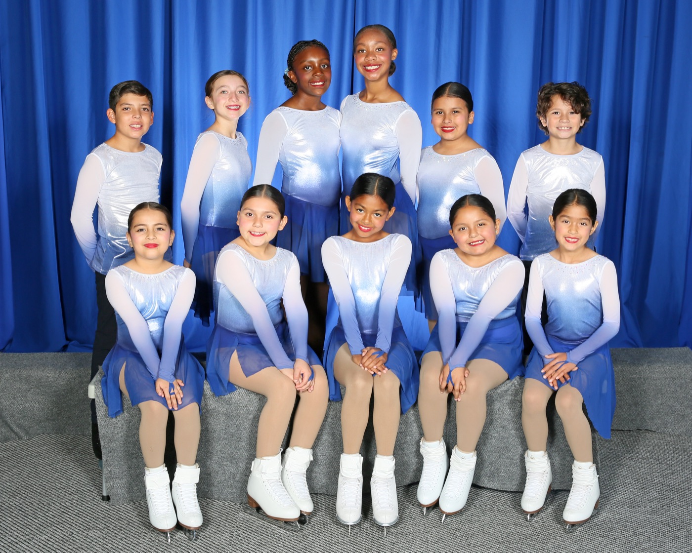 Team portrait of the eleven synchronized skaters in blue and white dresses in front of a blue curtain.