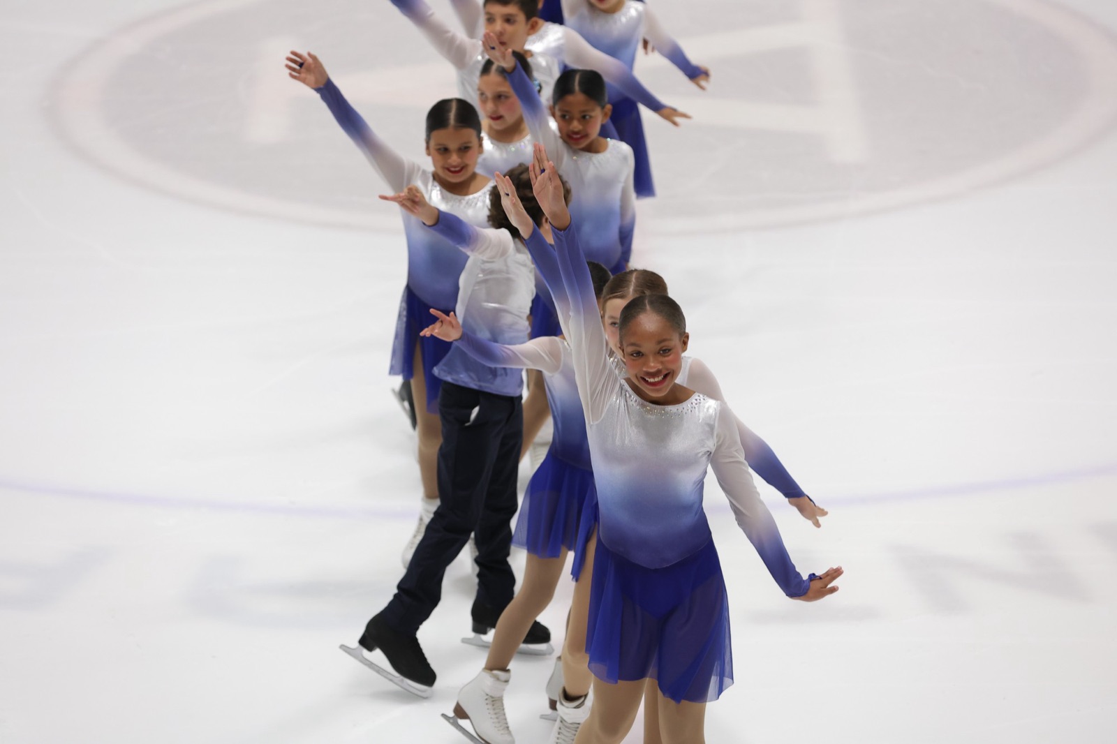 Synchronized skaters in blue and white dresses wave to the crowd as they glide in a single line, led by a smiling skater.